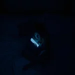 Person using smartphone in a dark bedroom before sleep, blue light emitting from screen.