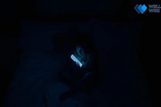 Person using smartphone in a dark bedroom before sleep, blue light emitting from screen.